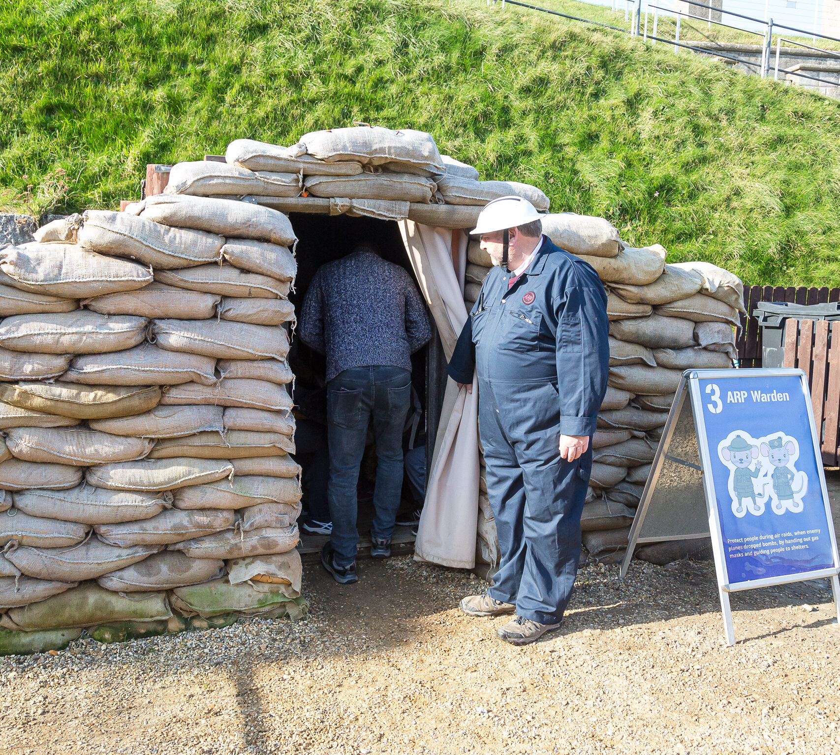 A man in a blue boiler suit is wear a black and white ARP warden tin hat in the style of WW2 era. He is holding a curtain back of the fort's anderson shelter, which is built from stacked sandbags and sheet metal. Behind the shelter is a grassy hill.