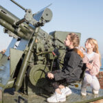Two young girls sit in the chair of the large green BOFUS gun that sits high on the ramparts.