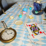 A photo of a table set up as the Mad Hatter's tea party - blue and white chequered table cloth with a large golden picket watch and an over-sized Queen of hearts card lay on the table, along with several cups and saucers, and a blue fabric top-hat.