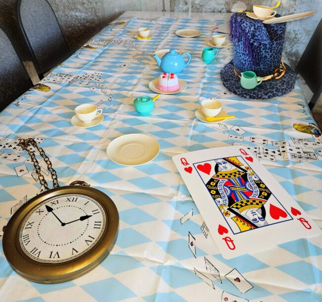 A photo of a table set up as the Mad Hatter's tea party - blue and white chequered table cloth with a large golden picket watch and an over-sized Queen of hearts card lay on the table, along with several cups and saucers, and a blue fabric top-hat.