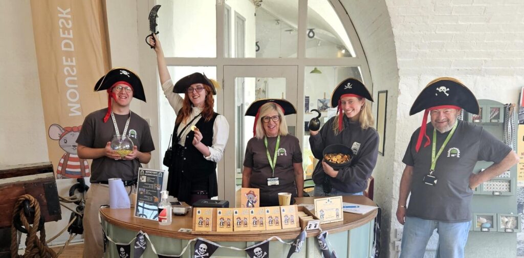 A group of volunteers and staff dressed as pirates for a pirate themed weekend. Most are wearing black pirate hats, and are standing behind the circular Mouse Desk which is covered in Pirate Jolly Roger bunting.