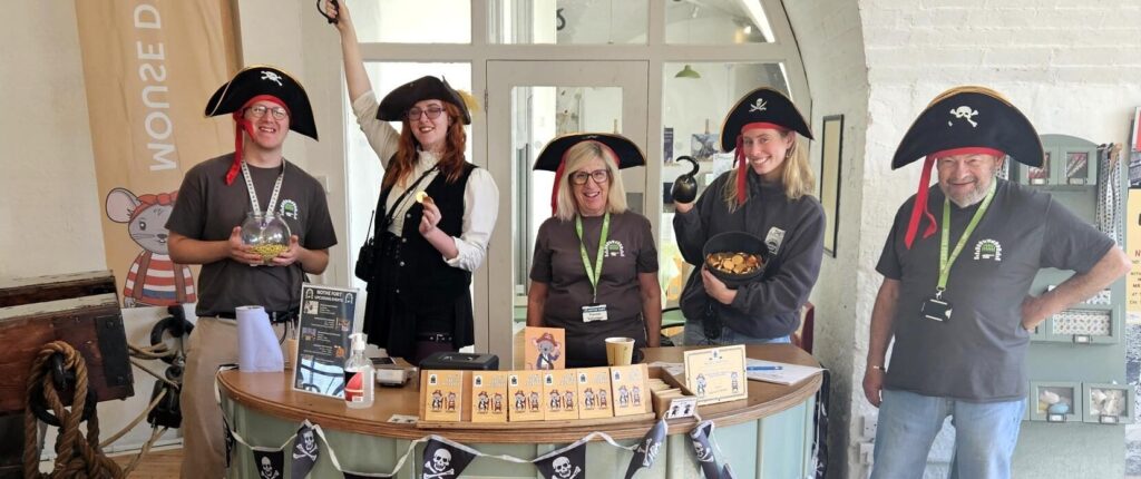 A group of volunteers and staff dressed as pirates for a pirate themed weekend. Most are wearing black pirate hats, and are standing behind the circular Mouse Desk which is covered in Pirate Jolly Roger bunting.