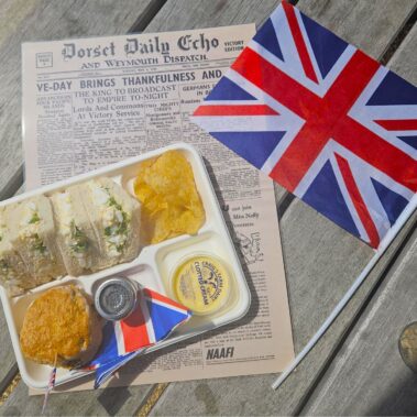 An overhead view of a takeaway picnic lunch to celebrate VE Day, with a tray of egg and cress triangle-cut sandwiches, a scone with jam and cream, and crisps. Under the tray is a VE Day newspaper and a union jack flag.