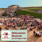 A photo of an outdoor performance in the Fort parade ground. There are many seats in rows in front of a stage where a group of 15+ performers are singing. There is a lovely clear blue sky evening behind the walls of the Fort.