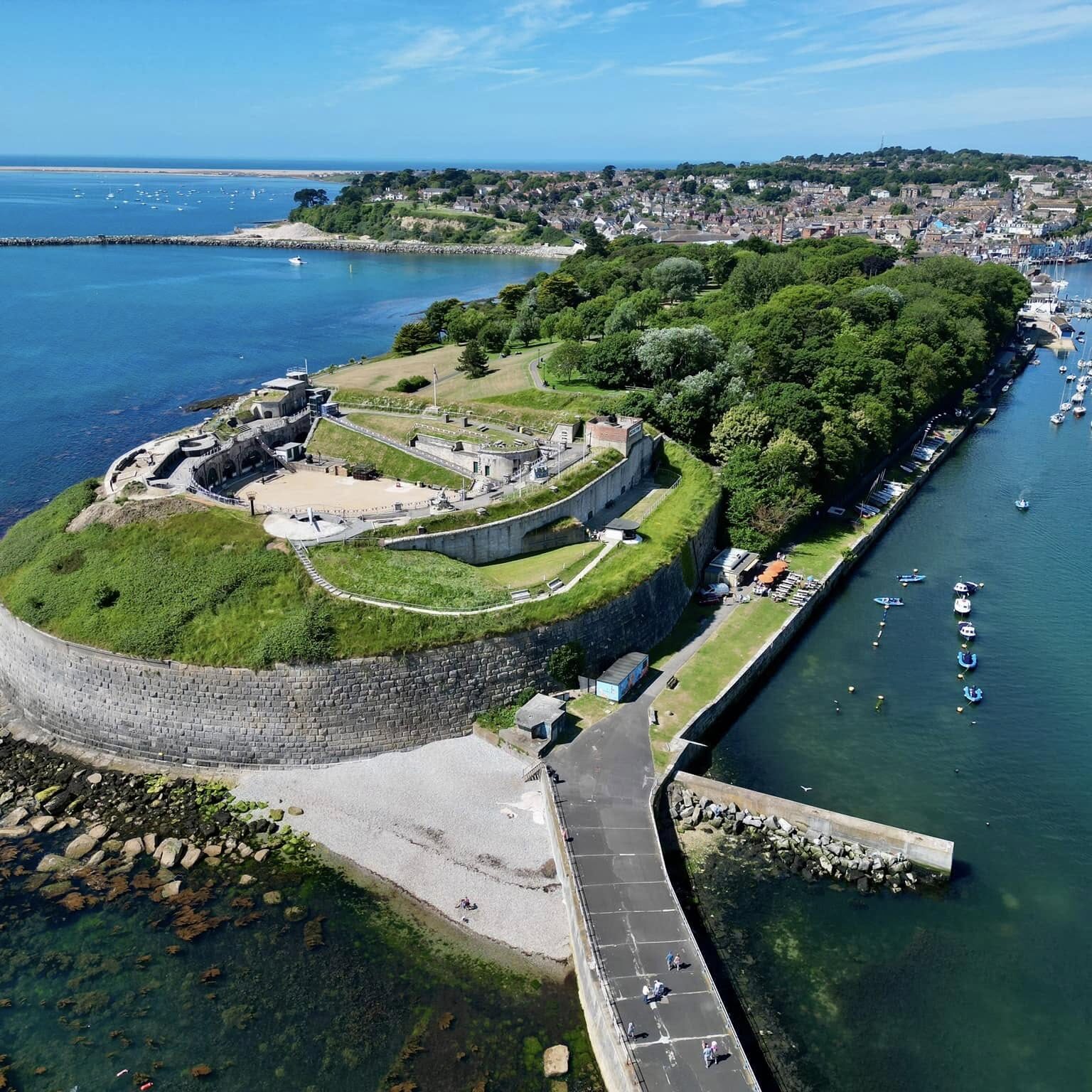 An aerial photo of Nothe Fort, with a clear blue sky above and the sea beyond contrasting with the bright green verges of the D shaped Fort.