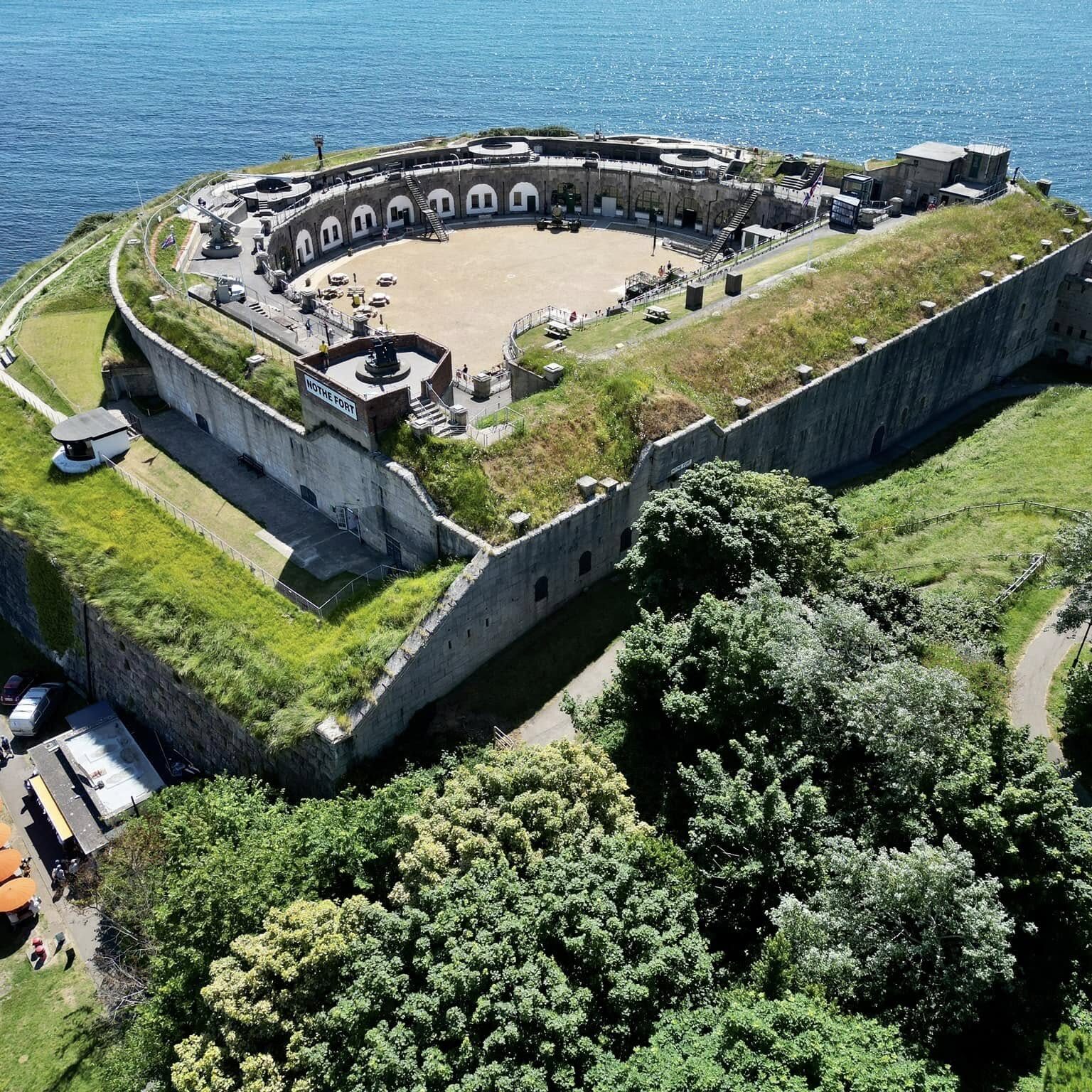 An aerial photo of Nothe Fort, with a clear blue sky above and the sea beyond contrasting with the bright green verges of the D shaped Fort.