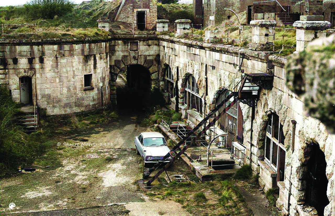 A photo taken in the 1970's of the abandoned Nothe Fort. Grass and overgrown weeds cover many of the stone walls, and there is a car parked below the stairs that lead from the mucky parade ground up to the overgrown ramparts.