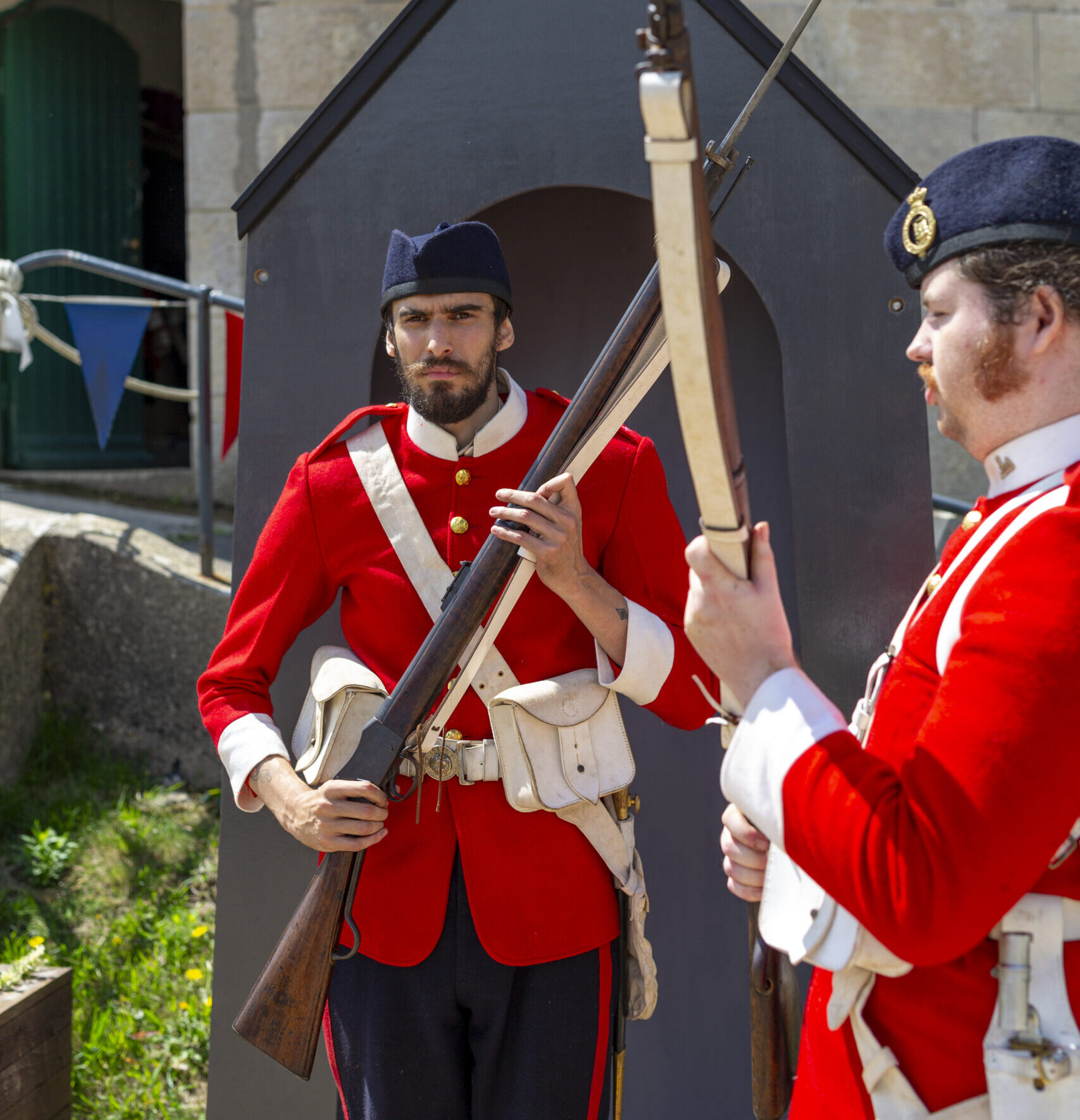 Two Victorian soldier reenactors from the Artillery Garrison stand in the parade ground outside a pillbox. Their red and white uniforms are vibrant in the sunshine.