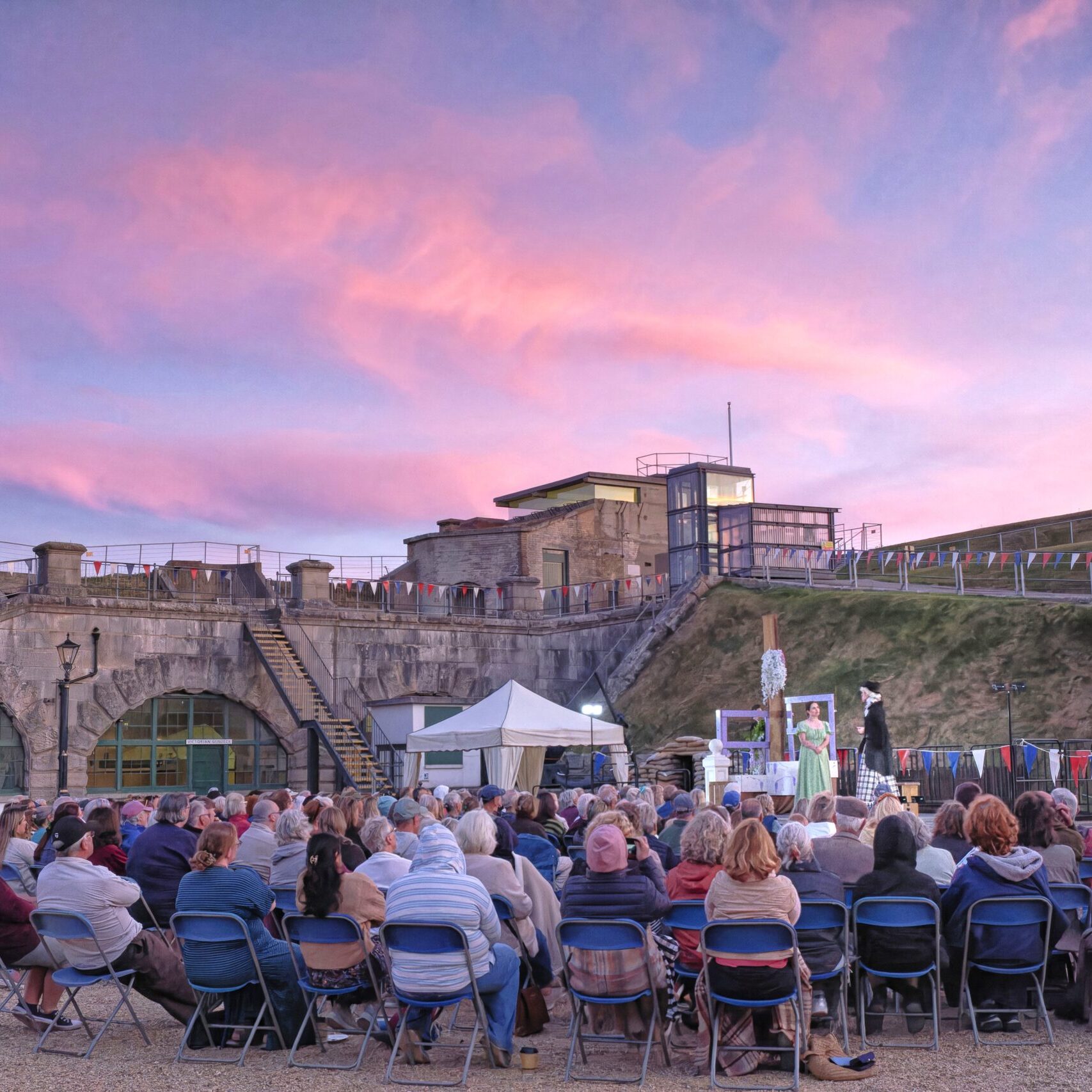 An outdoor evening theatre performance in the fort. The evening sky is a pretty pink and purple sunset. Many people are seated in front of the stage.