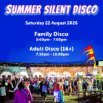 A photo of a previous silent disco at dusk in the parade ground of Nothe Fort. The stone walls are lit up with orange lights and in the centre of the image, a crowd of people are dancing and partying, waving glow sticks and wearing glowing headphones. At the top of the image, across a deep blue night sky, there is a glowy holographic looking font that reads "SUMMER SILENT DISCO Saturday 22 August"