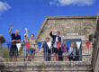 A family of visitors wave from up on the Fort ramparts. Behind them is a blue sky.