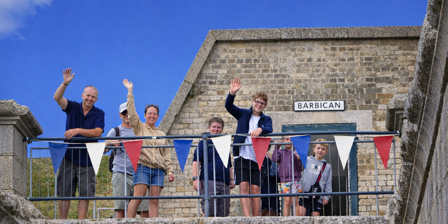 A family of visitors wave from up on the Fort ramparts. Behind them is a blue sky.