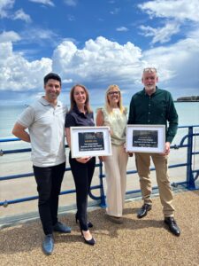 Photo of two Nothe Fort staff with two from South Coast embroidery holding two awards in frames: Nothe Fort Business of the Year for Weymouth, and the other group holds the Portland award. The photo is taken on the seafront of weymouth beach with Nothe Fort in the background and beautiful blue sky with fluffy white clouds.
