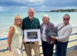 Photo of four Nothe Fort volunteers and staff holding two awards in frames: Business of the Year, and Beaumont Jones Community Service Award. The photo is taken on the seafront of weymouth beach with Nothe Fort in the background and beautiful blue sky with fluffy white clouds.