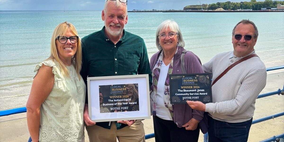 WBA 2026 double award Photo of four Nothe Fort volunteers and staff holding two awards in frames: Business of the Year, and Beaumont Jones Community Service Award. The photo is taken on the seafront of weymouth beach with Nothe Fort in the background and beautiful blue sky with fluffy white clouds.