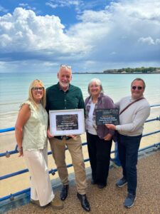 Photo of four Nothe Fort volunteers and staff holding two awards in frames: Business of the Year, and Beaumont Jones Community Service Award. The photo is taken on the seafront of weymouth beach with Nothe Fort in the background and beautiful blue sky with fluffy white clouds.