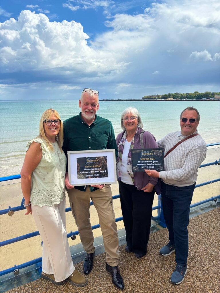 Photo of four Nothe Fort volunteers and staff holding two awards in frames: Business of the Year, and Beaumont Jones Community Service Award. The photo is taken on the seafront of weymouth beach with Nothe Fort in the background and beautiful blue sky with fluffy white clouds.