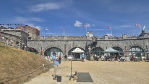 A photo of Paul Wise playing a bugle in the Nothe Fort Parade Ground with the iconic stone walls and union jacks flying against a blue sky. 