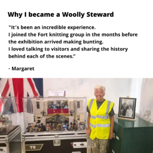 A photo of Margaret, a steward in a hi-viz yellow jacket standing beside their favourite knitted scene of Buckingham Palace. The text reads ' why i became a woolly steward: "It’s been an incredible experience. I joined the Fort knitting group in the months before the exhibition arrived making bunting. I loved talking to visitors and sharing the history behind each of the scenes.”