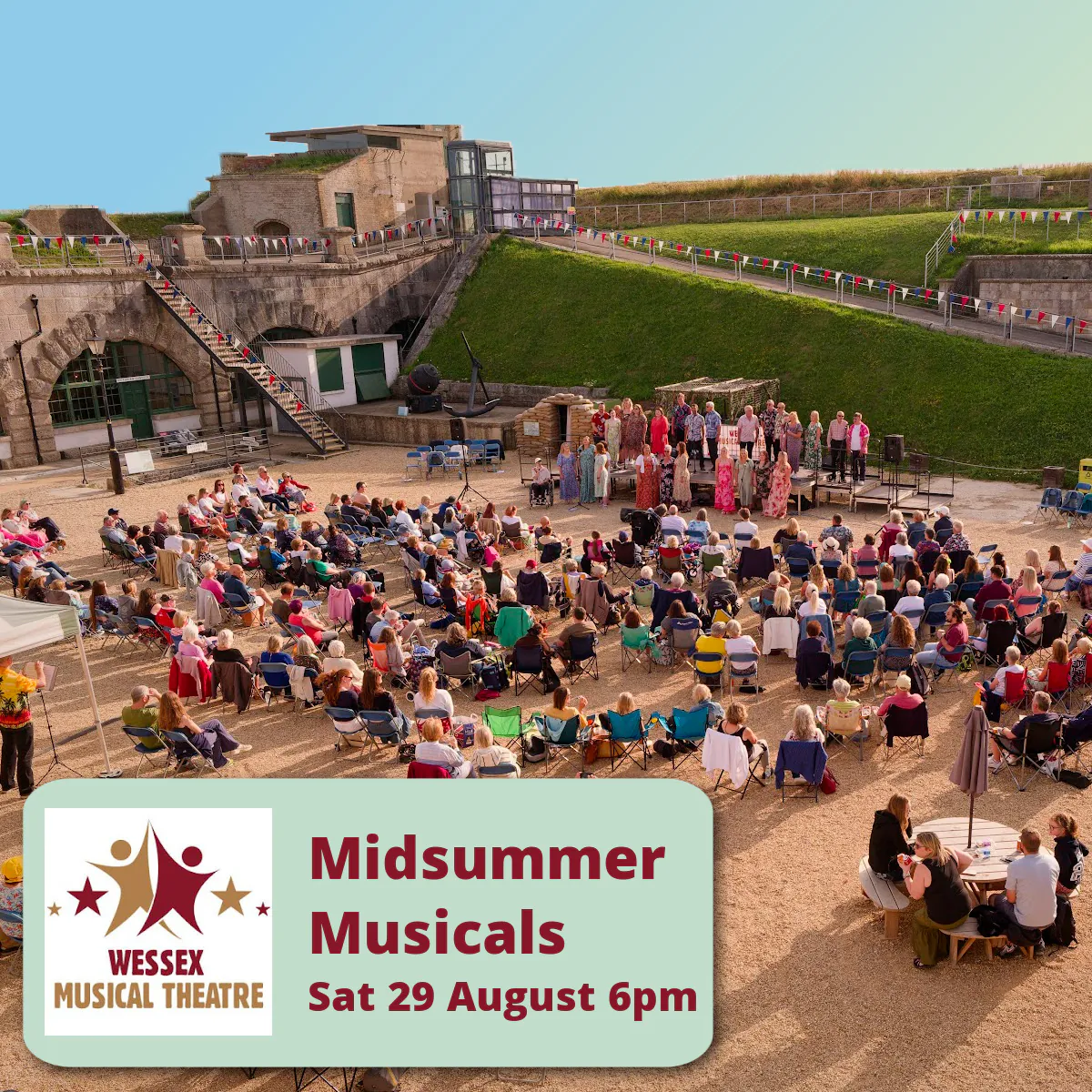 A photo of an outdoor performance in the Fort parade ground. There are many seats in rows in front of a stage where a group of 15  performers are singing. There is a lovely clear blue sky evening behind the walls of the Fort.